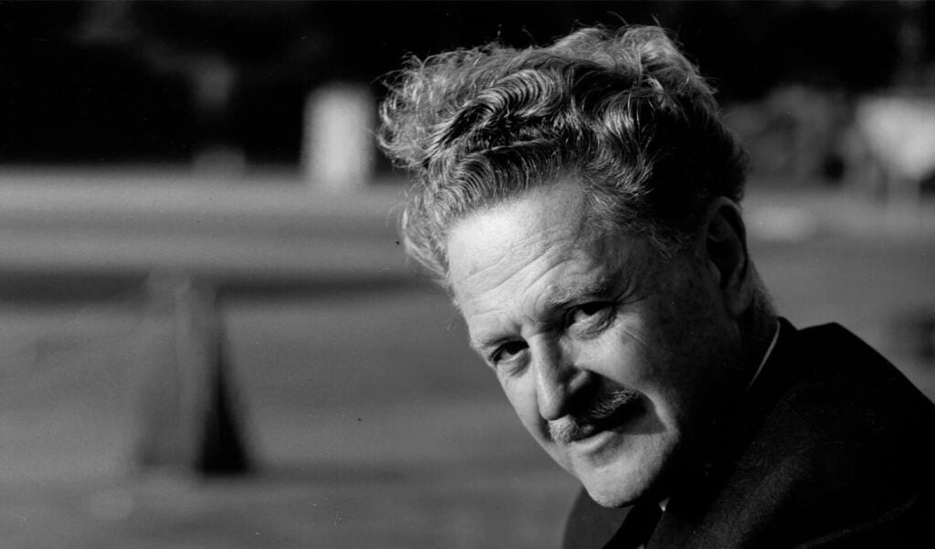 Monochrome close-up portrait of a man with wavy hair and a mustache, outdoors with a blurred background.