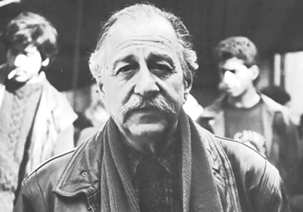 Older man with gray hair and a mustache, wearing a leather jacket and scarf, looking at the camera with blurred people in the background.