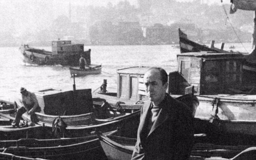 Man in a suit stands on a dock with small boats moored in the background.