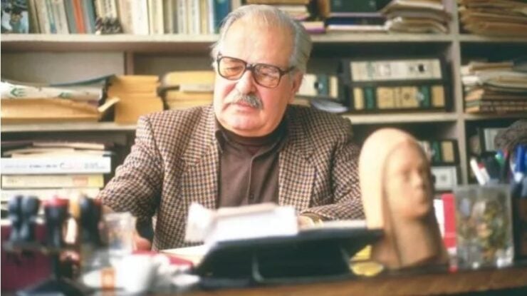 Older man with glasses and a mustache sits at a cluttered desk in a study, surrounded by books, looking at papers.