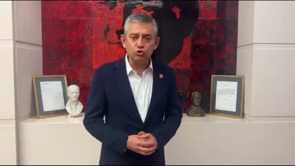 Man in a navy suit speaks to the camera in a room with red abstract art and framed documents behind him.