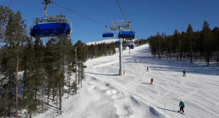 Blue ski chairlift cabins glide above a snowy slope with skiers and pine trees on both sides under a clear sky