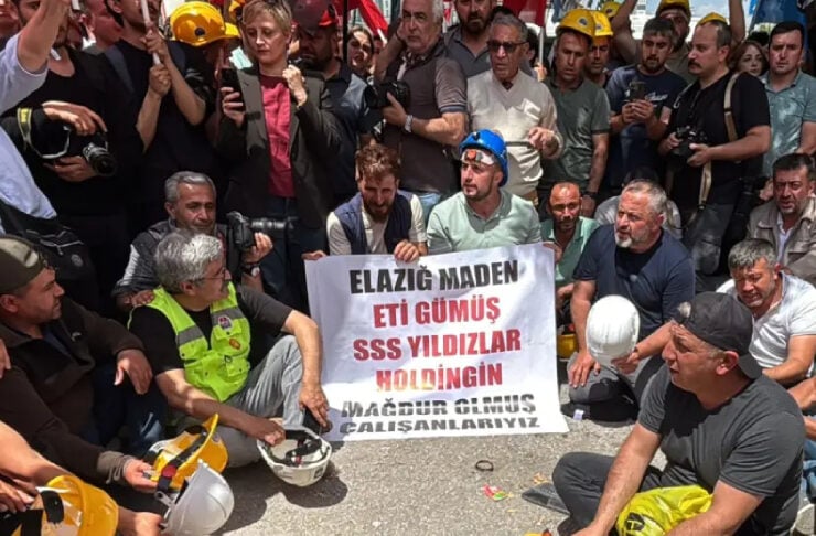 A crowd of miners and protesters sit and stand around on a street, holding a banner about Elazığ Maden and wearing hard hats.