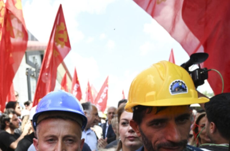 Two men wearing hard hats (blue on the left, yellow on the right) stand among a crowd at a protest with red flags in the background (construction/industrial workers vibe).