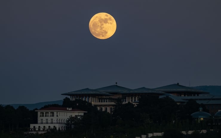 Full yellow moon rising over a dark city skyline at dusk, with trees and low buildings in the foreground.