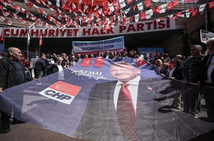 Crowd gathers at a Cumhuriyet Halk Partisi rally, holding a large banner with a portrait of a suited man and CHP logo nearby, red banners overhead.