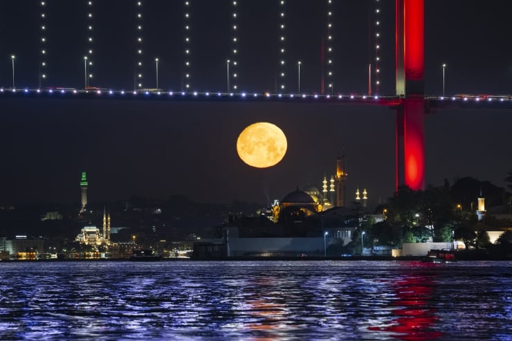 Night scene of a large orange moon rising over a city waterfront, with a lit bridge above and reflections on the water below.
