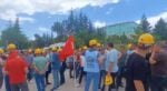 Group of construction workers wearing yellow helmets gather outdoors around a red flag during a break or protest.