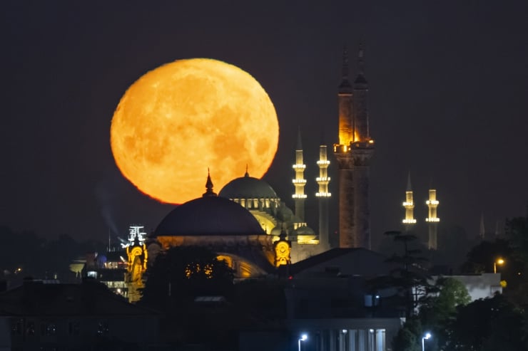 Large orange full moon rising behind a domed mosque with illuminated minarets at night.
