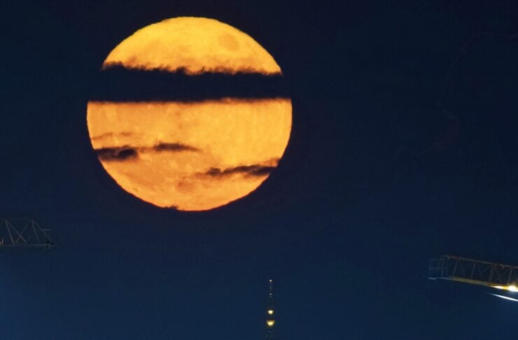 A bright orange full moon with dark atmospheric bands rising above a church spire against a night sky.