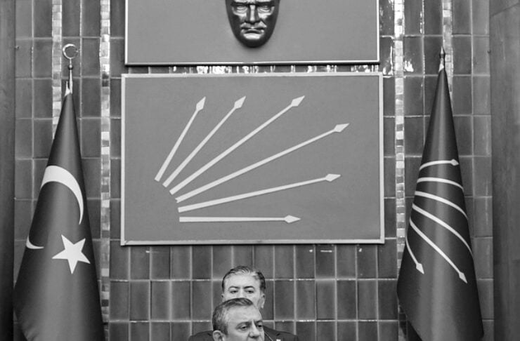 Male politician at a podium delivering a speech in a formal chamber, with two flags (left flag with star and crescent, right flag with curved lines) and a large emblem behind him and a panelist seated above.