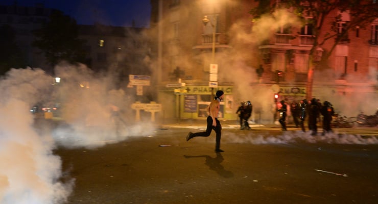 A shirtless person runs across a smoke-filled street at night as police in riot gear confront a crowd in the background.
