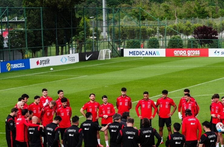 Turkish national team players in red training tops stand in a circle on a green soccer field listening to coaches in black jackets.