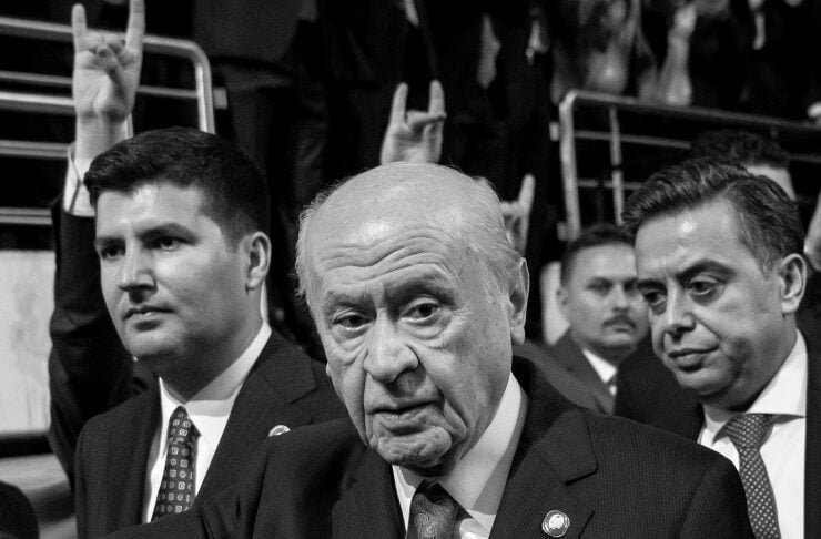 Older man in a dark suit and patterned tie at a formal event, with others in suits around him and audience members making rock-on hand gestures in the background (black-and-white photo).