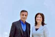 Two adults stand side by side, smiling; man wears a dark suit with a blue shirt, woman wears a light gray sweater against a pale blue wall.