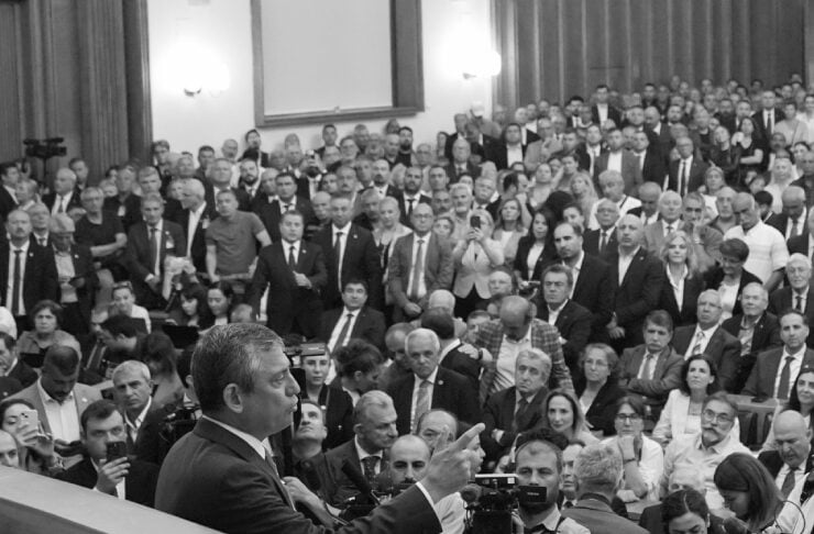 Speaker at a podium addressing a large seated crowd in a formal gathering, with journalists and cameras nearby
