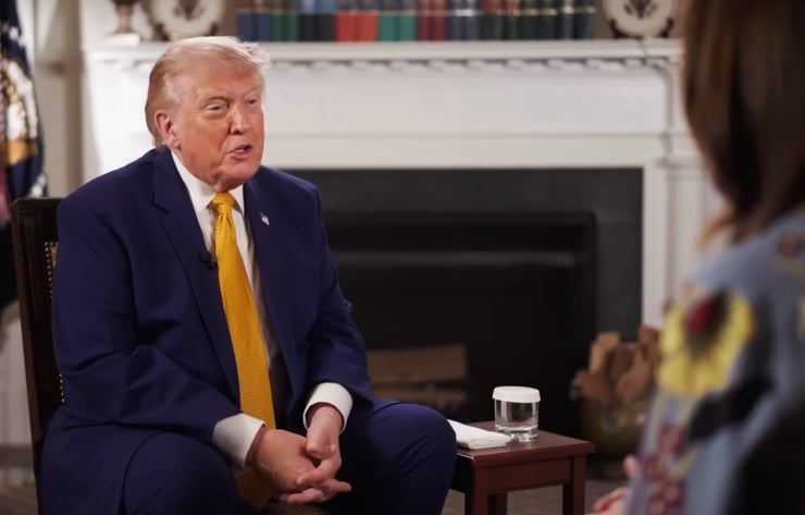Former President Donald Trump in a navy suit and yellow tie speaks during an interview, seated in a formal office with a glass of water on a side table and a fireplace in the background.