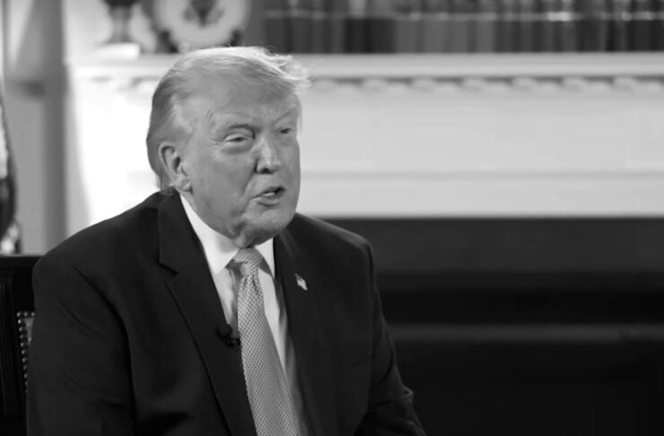 Grayscale photo of a man in a dark suit and tie speaking during an interview, hands clasped, with a glass of water nearby.