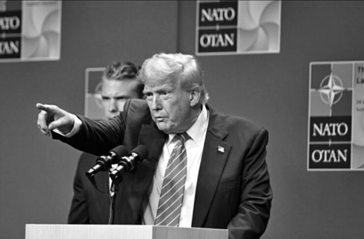 Donald Trump at a podium with NATO logos, pointing and speaking into microphones during a press conference; backdrop shows 'NATO OTAN' signs.