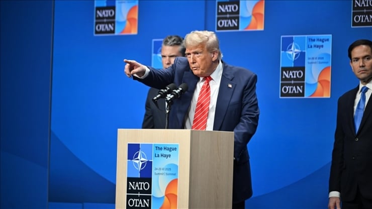Donald Trump at a NATO press conference, pointing with his right hand from a podium with NATO banners in the blue backdrop.