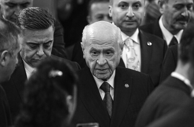 Older man in a dark suit at a formal gathering, surrounded by people, looking down