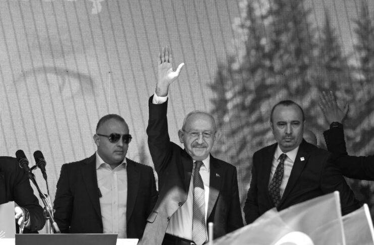 Man in a suit raises his right hand on a stage, flanked by security, as a crowd cheers with Turkish flags nearby.