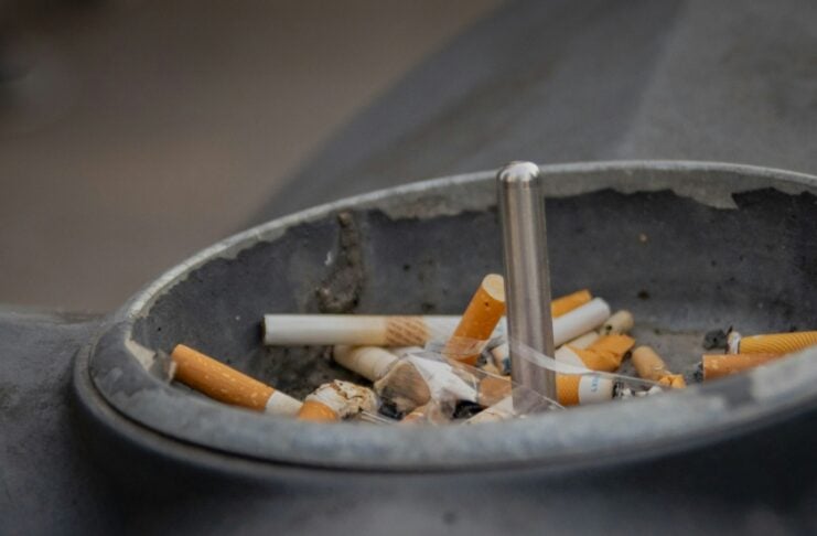 Close-up of a crowded ashtray filled with cigarette butts and a metal cigarette holder in the center, showing smoke nearby.