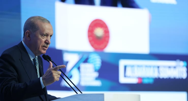 Male political leader delivering a speech at a podium with a Turkish flag displayed on a large screen behind him behind the podium.