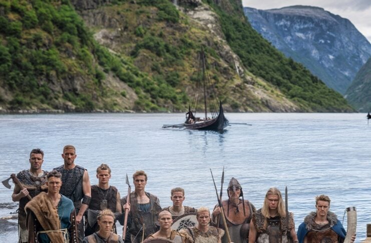 Group of Viking reenactors posing on a rocky shore with round shields and weapons, a longboat on calm water behind them and green cliffs in the background.