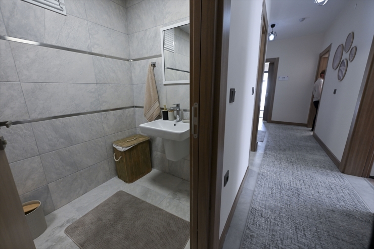 Gray-tiled bathroom with a white sink, mirror, towel, and a wicker laundry basket beside the sink, visible from an open door into a hallway.