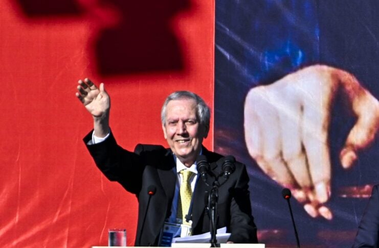 Man in a black suit and yellow tie speaks at a podium with the Fenerbahçe logo, raising his right hand.