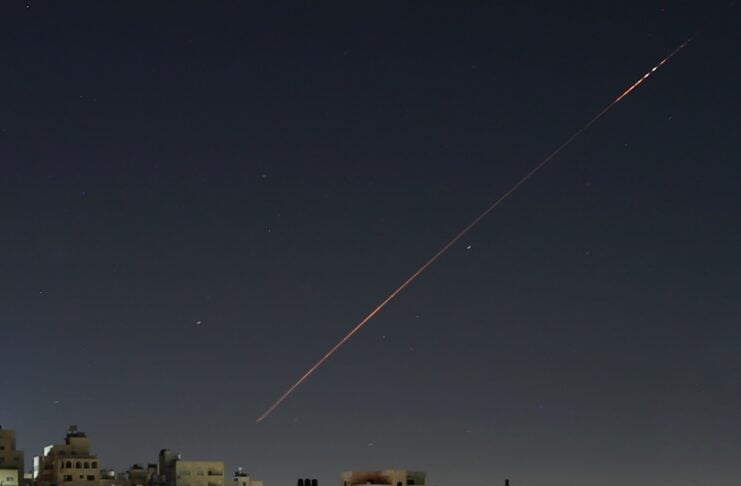Night city skyline at night with a red-orange diagonal rocket trail across the sky, from the urban horizon to the upper right corner.