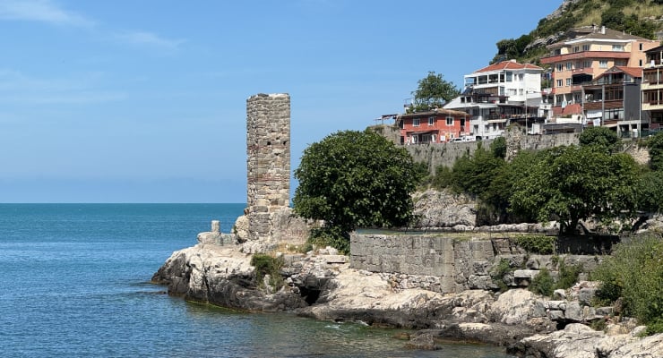 Stone sea wall with a tall, ancient cylindrical tower ruin overlooking a rocky shoreline and calm blue sea; colorful houses on a hillside in the background.