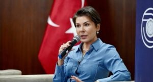 Woman in a blue shirt speaks into a handheld microphone at a panel, with a Turkish flag in the background.