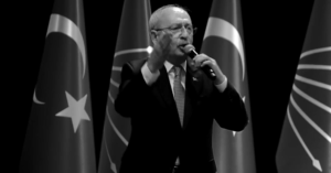 Male speaker in a suit on stage, holding a microphone, with Turkish flags behind him on either side.