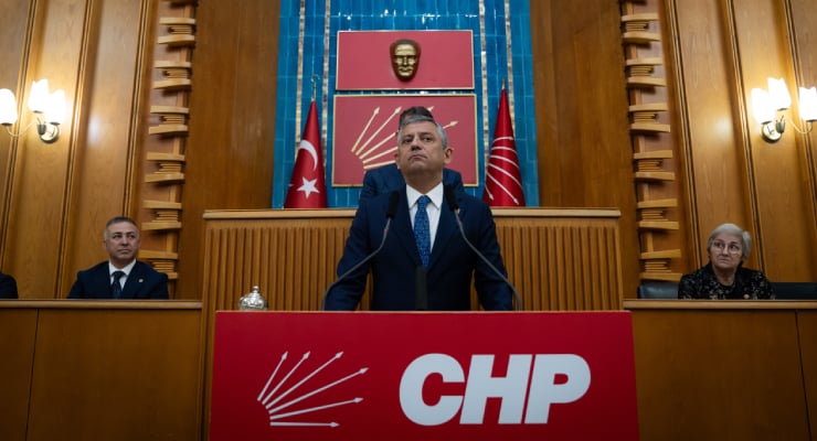 Politician in suit delivers speech at a wooden podium in a Turkish parliament chamber, with CHP banner and flags behind him.