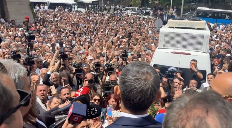 Crowd of reporters and bystanders surround a man in a suit giving a speech outdoors; numerous cameras, microphones, and smartphones aimed at him, with a white vehicle in the background.