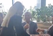 Woman in a black jacket hands a rectangular gift box to a seated man during a casual gathering.