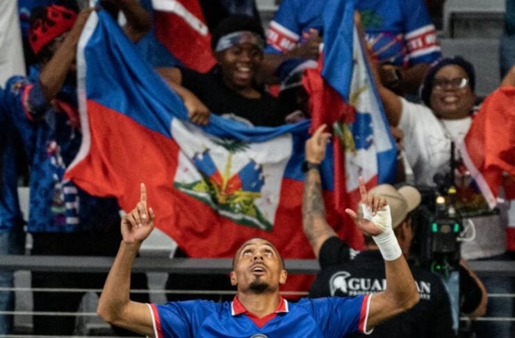 Male player in blue jersey 18 raises both arms in celebration as fans wave flags behind him in the stands.