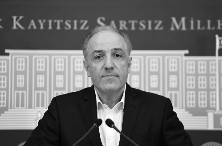 Man in a dark suit speaks at a press conference, standing behind two microphones with a government-style backdrop behind him.