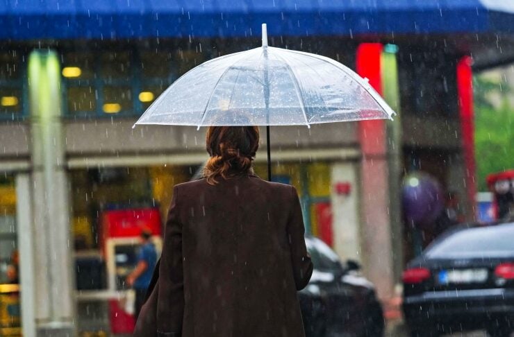 Person seen from behind walking in the rain, holding a clear umbrella in a city street; storefronts blur in the background.
