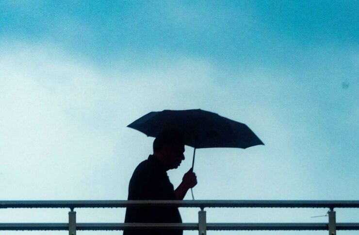 Person walks along a railing while holding an umbrella against a cloudy blue sky, silhouette in profile.