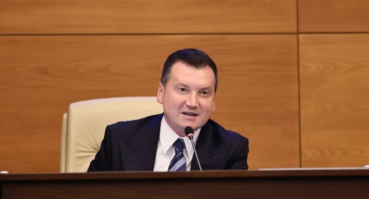 Man in a dark suit and striped tie speaks into a microphone at a wooden podium in a formal meeting room about to address an audience.