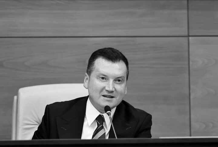 Man in a suit speaks into a microphone at a formal podium in a wooden-paneled setting