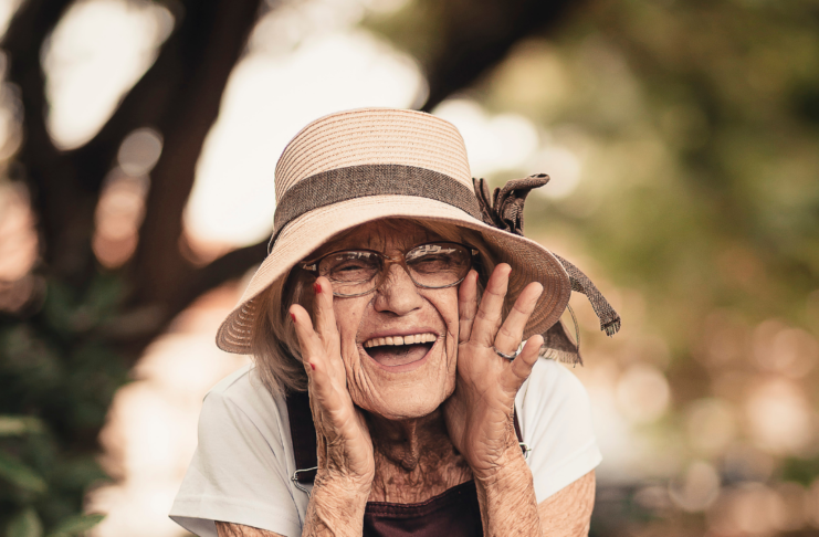 Smiling elderly woman wearing a straw hat and glasses, cupping her cheeks with both hands outdoors.