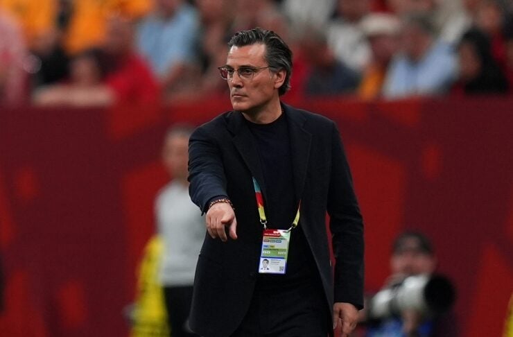 A man in a dark suit with a lanyard and badge points toward the field during a sports event on a red-background stadium.