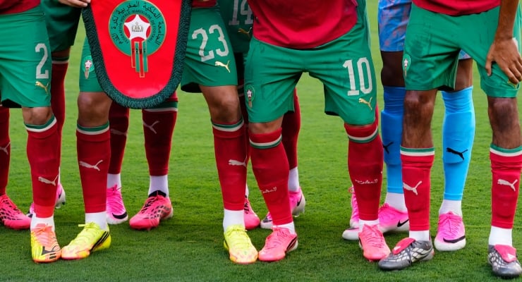 Soccer players standing in a line on a green field, wearing red jerseys and green shorts with colorful cleats; a team banner is nearby.