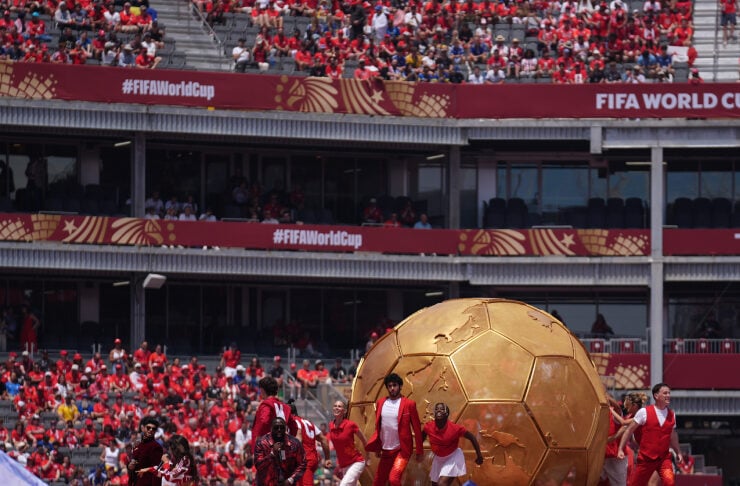Stage performance at a stadium with a giant golden soccer ball prop, dancers in red and white, and a cheering red-clad crowd under FIFA World Cup banners.