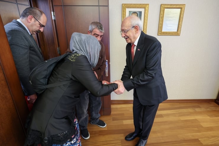 A man in a dark suit shakes hands with a woman wearing a headscarf in a doorway, while two other men look on in a hallway office setting.