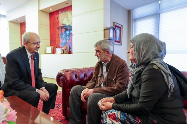 Three adults seated in a reception area, a man in a suit talking with a bearded man and a woman wearing a headscarf.